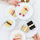 Small cakes on white plates with hands reaching for them on a light background